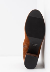 Bruine suède enkellaars met een lage gestapelde hak, een getextureerde zool en zijritssluiting. Maat 37 staat op de zool vermeld.