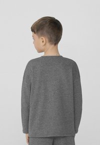 Šedá dlouhá košile s dlouhým rukávem vyrobená z měkké tkaniny, s kulatým výstřihem a uvolněným střihem, zobrazená na dítěti zezadu.