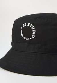 Mustapucket-hattu, joka on valmistettu teksturoidusta kankaasta ja jossa on pyöreä valkoinen logo, jossa lukee "JJ STUDIO" ja "DENIM CREW". Vaakasuora nauhadesign.
