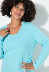 Hellblaues Langarmshirt aus weichem Stoff mit weitem Halsausschnitt und lockerem Schnitt. Das Modell hat lockiges Haar und trägt schlichte Ohrringe.