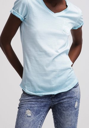 Osoba ubrana w jasnoniebieski, krótki rękaw, rozpinany pod szyją T-shirt z kieszenią i podwiniętymi mankietami, w połączeniu z niebieskimi, podartymi jeansami, z rękami za plecami.