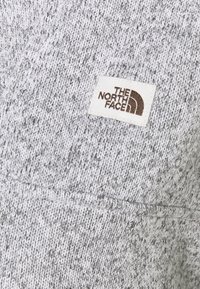 Grauer Strickpullover mit einem strukturierten Muster; verfügt über ein weißes Etikett mit dem braunen "The North Face"-Logo, das auf den Stoff gestickt ist.