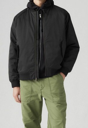 Veste bomber noire avec capuche, dotée d'une fermeture éclair et de poches latérales. Assortie à un pantalon cargo vert clair. Texture de tissu lisse.