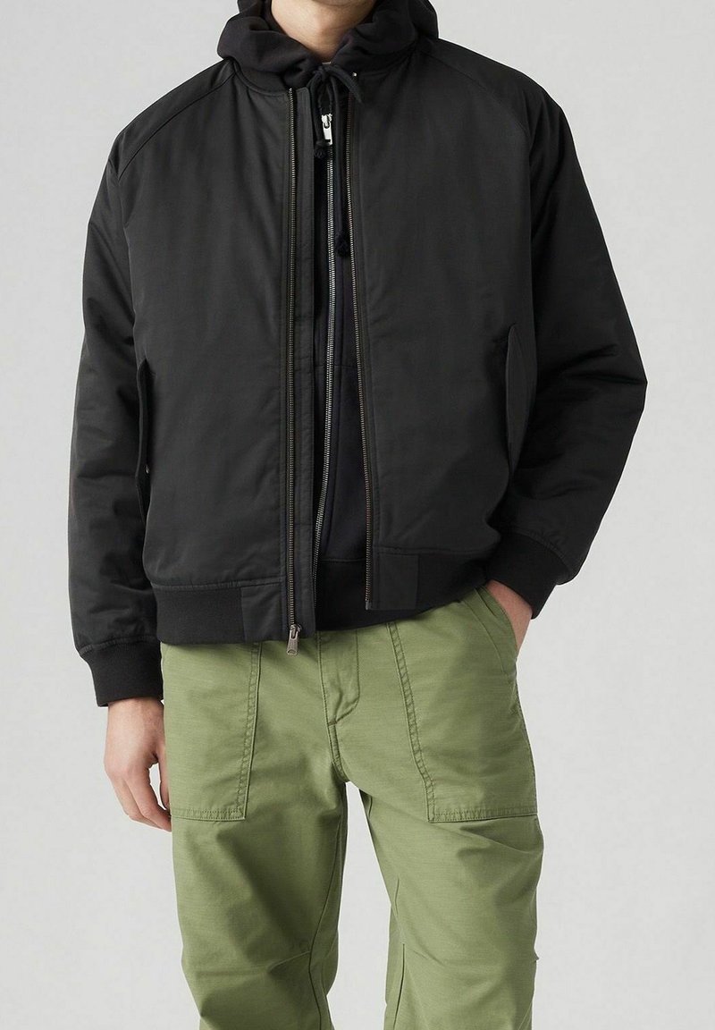 Svart bomberjacka med huva, med dragkedjestängning och sidofickor. Kombinerad med ljusgröna cargobyxor. Slät tygtextur.