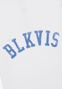 T-shirt blanc avec des lettres bleuissantes légèrement usées épelant "BLKVIS" dans un design arqué sur le devant.