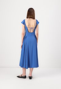 Robe sans manches bleue avec un dos plongeant et un détail à nouer. Tissu lisse, jupe fluide et manches volantées. Portée avec des chaussures plates noires.