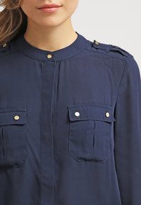 Camicia abbottonata blu navy con scollatura tonda, dettagli in bottoni color oro e due tasche frontali con pattina. Tessuto dalla texture liscia.