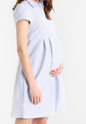 Femme enceinte portant une robe à manches courtes rayée bleu clair, tenant doucement son ventre, debout devant un fond blanc.