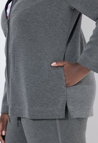 Veste zippée grise avec poche latérale, texture douce, manches longues et ourlet fendu. Associée à un pantalon gris assorti avec un détail de couture.