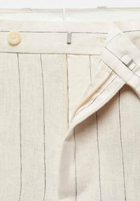 Pantalon en lin beige avec fines rayures verticales noires, fermeture à crochet et barre en métal, et un petit bouton beige sur la taille.