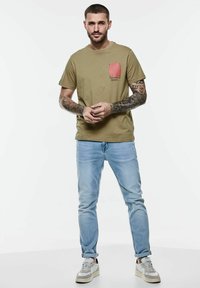 Camiseta de manga corta verde olivo con un estampado de bolsillo gráfico rojo, combinada con jeans azul claro y zapatillas claras con detalles en gris.