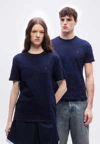 Νέος άνδρας και γυναίκα που φορούν μπλε ναυτικό μπλουζάκια με μικρό λογότυπο, στέκονται δίπλα-δίπλα μπροστά σε απλό ανοιχτό φόντο.