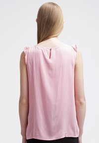 Helles rosa, ärmelloses Blusenshirt aus glattem Stoff. Mit Rüschendetails an den Schultern und einer kleinen Schlüssellochöffnung auf der Rückseite.