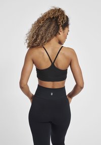 Vrouw met krullend haar, die een zwarte sportbh met bandjes en high-waisted leggings draagt, staat met handen in de zij, met de rug naar de camera.