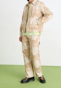 Beige vzorovaný jacket a kalhoty vyrobené z bavlněné směsi, s blokovými vzory, v kombinaci se světle zeleným topem a černými robustními botami.