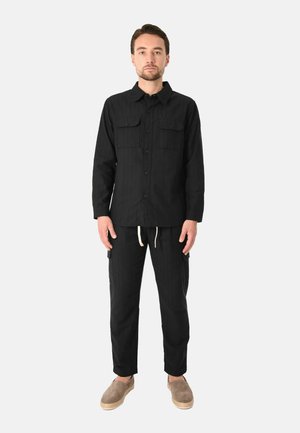 Uomo in piedi che indossa una camicia nera con bottoni e tasche sul petto, pantaloni neri con coulisse e scarpe marroni slip-on su sfondo bianco.
