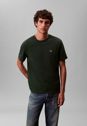 Jonge man met krullend haar, gekleed in een donker groen T-shirt met het cK-logo en een blauwe spijkerbroek, staand tegen een effen grijze achtergrond.