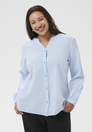 Usmievavá žena s dlhými tmavými vlasmi, oblečená v svetlomodrej košeli na gombíky a čiernych nohaviciach, stojaca pred jednoduchým pozadím.