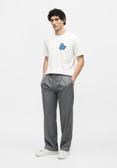 Νέος άντρας με σγουρά μαλλιά, φοράει λευκό μπλουζάκι με μπλε λαγουδάκι, γκρι πλισέ παντελόνια, λευκά αθλητικά παπούτσια, στέκεται με τα χέρια στις τσέπες.