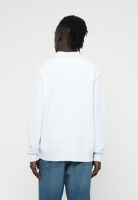 Ανοιχτό μπλε φλις πουλόβερ με βουρτσισμένη υφή, γιακά, ριμπ μανσέτες και άνετη γραμμή, φορεμένο με μπλε τζιν παντελόνι.