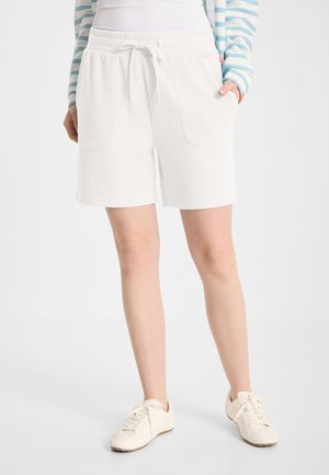 Persoon die witte shorts met trekkoord en grote voorkanten draagt, witte sneakers en een gestreept shirt met lange mouwen.
