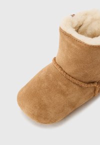 Tan mockasiner med en mjuk, krämfärgad pälsfoder. Har en rundad tå och dekorativ sömnad längs med ovandelen.