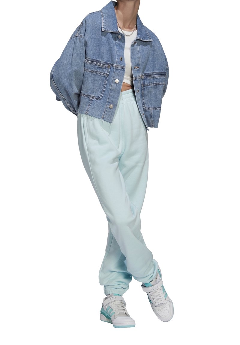 Osoba ubrana w skróconą niebieską dżinsową kurtkę, białą bluzkę crop top, jasnoniebieskie dresy i białe trampki z niebieskimi akcentami, stojąca z założonymi nogami.