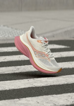 Chaussure de course blanche et rose en suspension au-dessus d'un passage pour piétons sur une route asphaltée, montrant les détails de la semelle et du côté.