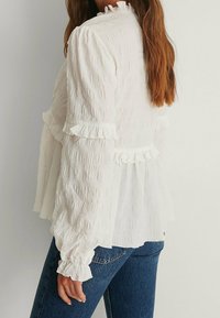 Blusa bianca in tessuto texturizzato con dettagli a volant sulle maniche e sull'orlo, caratterizzata da un colletto alto e una vestibilità ampia, abbinata a jeans blu scuri.