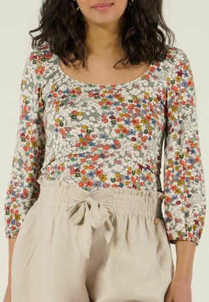 Vrouw draagt een langarmige top met bloemenprint in rood, geel en blauw, gecombineerd met hooggetailleerde beige broek met een strik aan de voorkant.