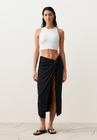 Vrouw draagt een wit mouwloos crop topje, een zwarte wikkel midi rok met een vooraan een split en zwarte teenslippers, staand op een lichte vloer.