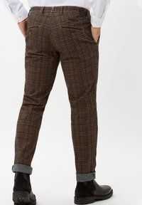 Homme portant un pantalon marron à carreaux avec des revers et des bottes en cuir noires, debout les mains dans les poches sur un fond blanc uni.