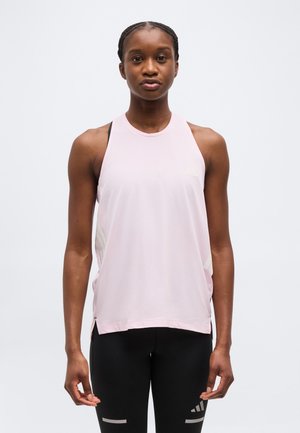 Mladá žena v svetloružovom športovom tielku bez rukávov a čiernych legínach stojaca pred jednoduchým svetlým pozadím.