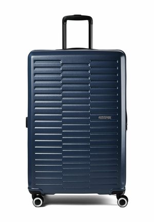 Valigia rigida blu navy con scanalature orizzontali, quattro ruote girevoli, manico telescopico e logo American Tourister sul davanti.