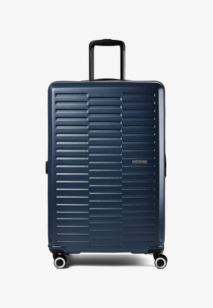Valigia rigida blu navy con scanalature orizzontali, quattro ruote girevoli, manico telescopico e logo American Tourister sul davanti.