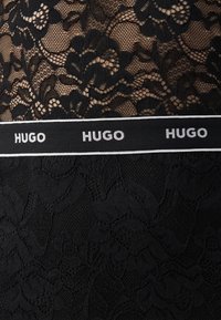 Černá krajka s květinovými vzory, s pevním černým pasem označeným "HUGO" v bílé barvě. Hladká textura s propracovanými detaily.