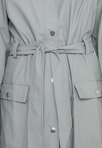 Jasnoszary płaszcz przeciwdeszczowy z paskiem w talii, zatrzaskami, dwoma przednimi kieszeniami oraz gładką, wodoodporną fakturą.