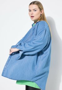 Camicia oversize in denim blu con bottoni e tasca, sovrapposta a un top verde. Presenta maniche lunghe e orlo curvo.
