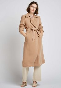 Trench beige clair avec un lien à la taille, deux poches latérales et un col rabattu. Associé à un pantalon large crème et des sandales beige à enfiler.
