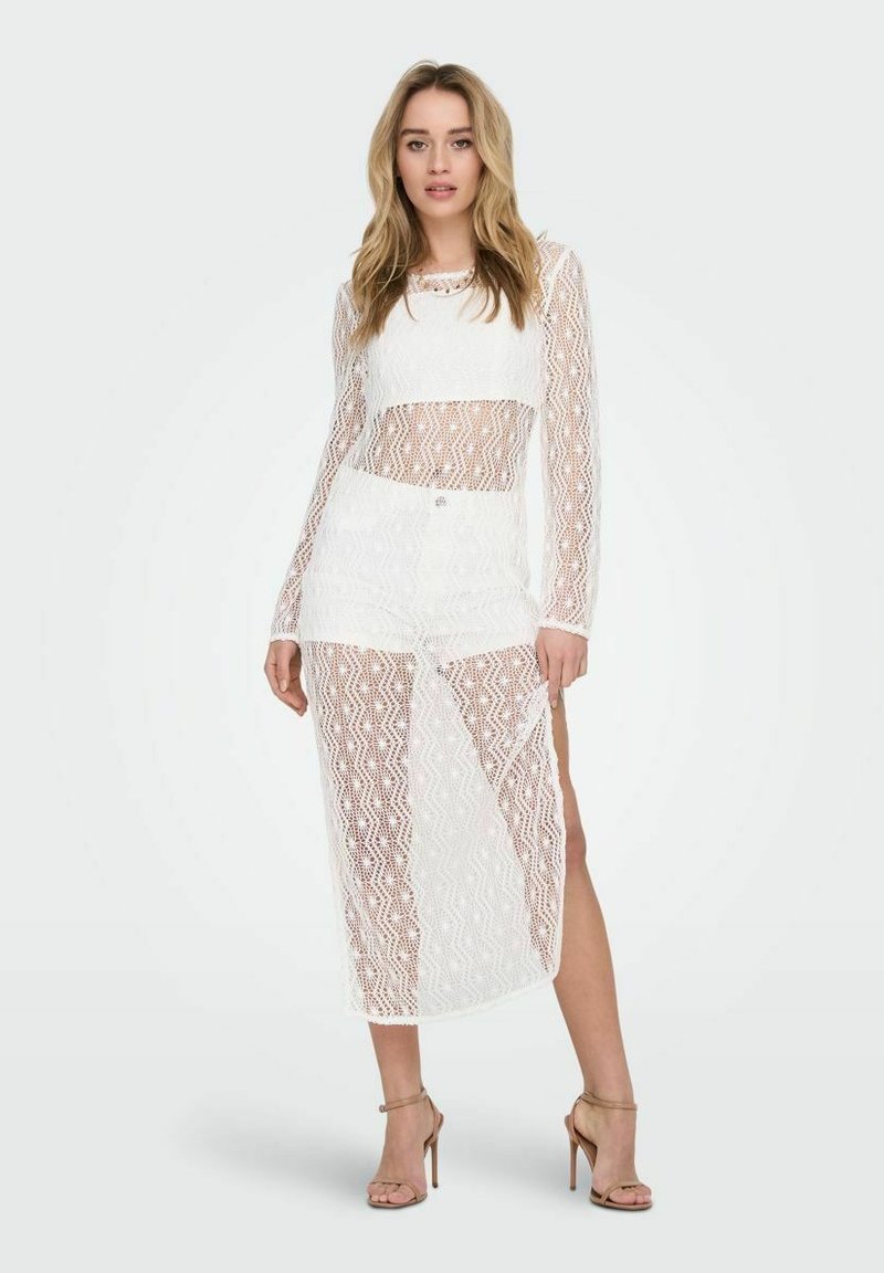 Weißes Spitzen-Zweiteiler-Outfit mit einem Langarmoberteil mit transparenten Einsätzen und einem passenden Rock, beide mit einem geometrischen Muster und einem Schlitz bis zur Mitte des Oberschenkels.