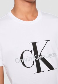 White crew neck t-shirt with large black "CK" and gray "Calvin Klein Jeans" logo worn by person shown from neck to chest.