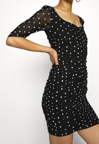 Musta rypistetty mini mekko valkoisilla pilkuilla ja läpinäkyvillä kolmannes hihoilla, käsi lantion päällä, ylävartalo näkyvissä.