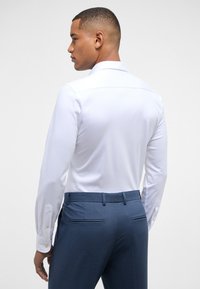Weißes Langarmhemd mit klassischem Kragen, kombiniert mit blauen maßgeschneiderten Hosen, die über Gesäßtaschen und Knöpfe an den Bündchen verfügen.