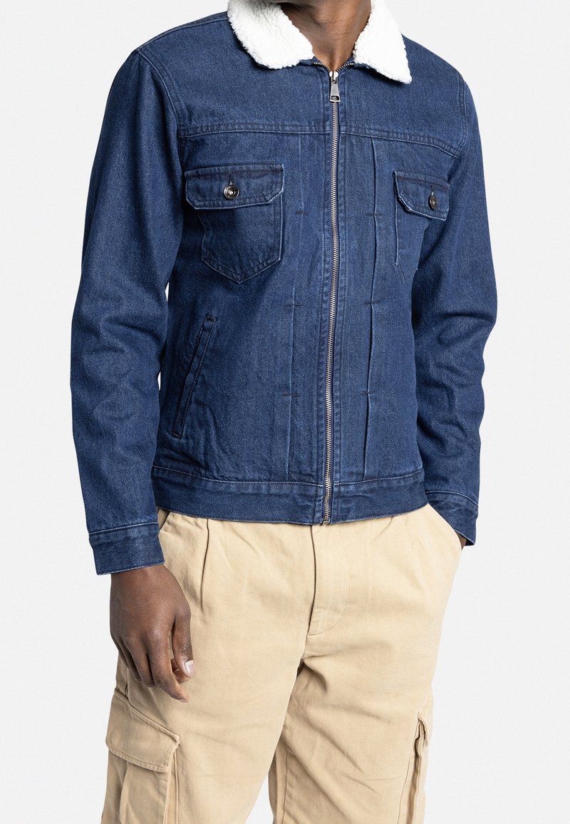 Veste en denim bleu foncé avec un col en fausse fourrure blanche, deux poches poitrine, fermeture éclair et poignets ajustés, associée à un pantalon cargo beige.