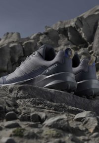 Σκούρα γκρι πεζοπορικά παπούτσια Adidas Terrex που ξεκουράζονται σε βραχώδες έδαφος κάτω από καθαρό ουρανό.