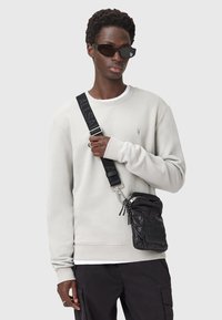 Felpa grigia chiara con polsini a coste, borsa nera con tracolla regolabile e cerniere, e accenti rettangolari neri. Pantaloni scuri e occhiali da sole.