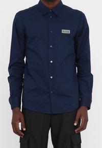 Camicia a maniche lunghe blu navy, con chiusura a bottoni, in tessuto intrecciato, con una patch del logo sul petto sinistro e bottoni blu.