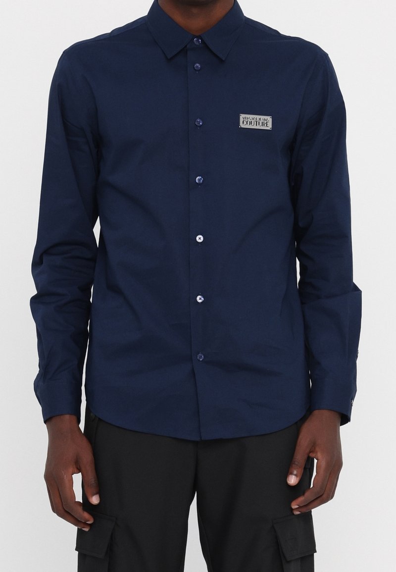 Camicia a maniche lunghe blu navy, con chiusura a bottoni, in tessuto intrecciato, con una patch del logo sul petto sinistro e bottoni blu.