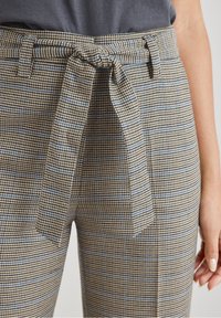 Pantalons à motifs à carreaux beige et bleu avec une ceinture en tissu à la taille. Le matériau semble texturé avec une coupe ajustée.