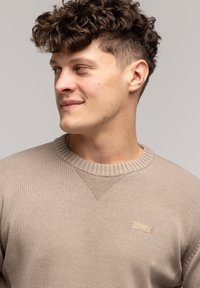 Beżowy sweter dzianinowy z okrągłym dekoltem i prążkowanymi mankietami, ozdobiony małą, prostokątną skórzaną metką na piersi z nazwą marki.